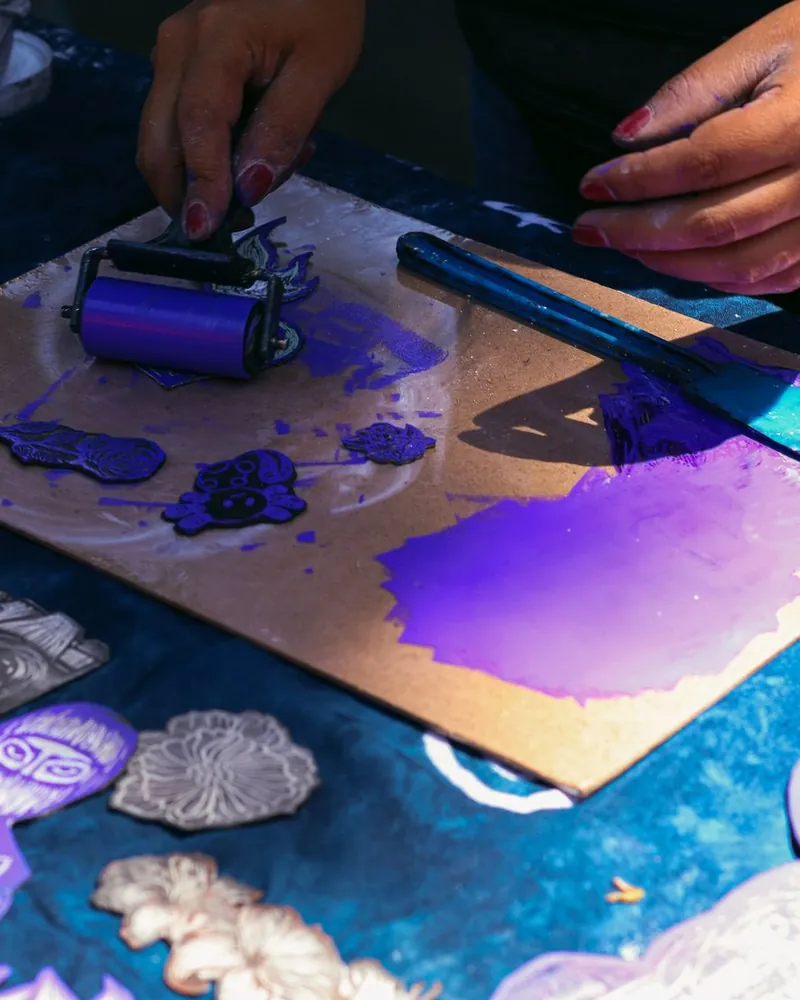 Rodillo de mano extendiendo tinta violeta sobre una plancha, junto a matrices de linóleo talladas y una espátula con tinta.